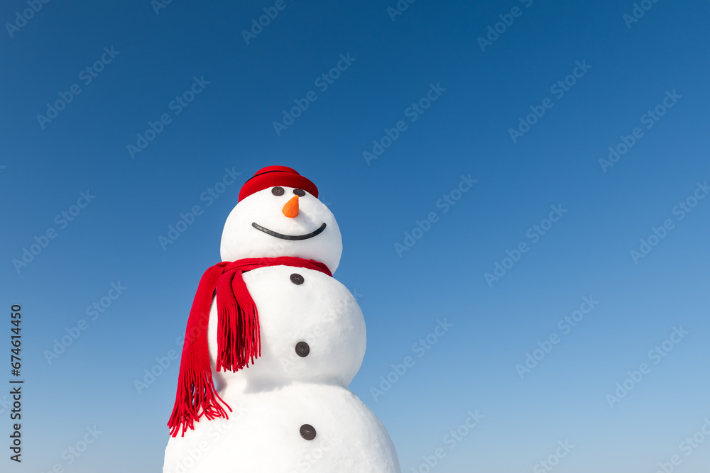 In a snowy landscape, a jolly snowman wears a stylish red hat and a matching red scarf, all under a vibrant blue sky. Christmas postcard