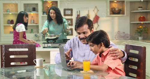Happy Indian family father teaching son to use laptop while mother and daughter preparing food in kitchen
