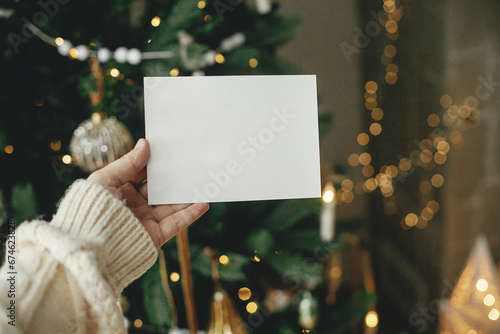 Christmas card mock up. Hand holding empty greeting card on background of stylish decorated christmas tree with golden lights. Space for text. Season greetings template and vintage ornaments