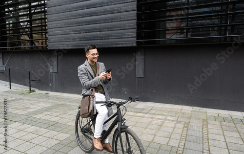 Happy employee environment lover calling colleagues before staff meeting. Cheerful businessman with glasses on reading text messages on a smart phone and pushing a bicycle.