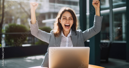 A happy young businesswoman celebrating success with arms raised in front of a laptop, fists clenched. The freelancer has finished a project