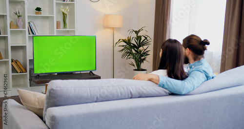 Fotografie Happy man and woman sitting at home in living room watching green chroma key screen television, relaxing on a sofa