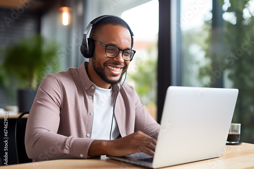 African male student wearing headphones and learning language on distance online at laptop. Sitting at desk at office. Black guy watching webinar, listening lecture. E-learning, remote education.