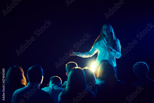 Young woman, stand up comic standing on stage, laughing, cheerfully telling audience funny stories and jokes. Concept of entertainment, fun and joy, concert, performance, show