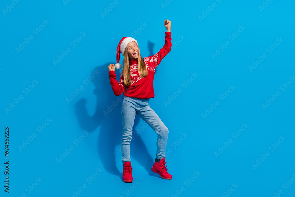 Full size photo of overjoyed schoolgirl dressed red sweater raising fists up celebrate christmas isolated on blue color background Full size photo of overjoyed schoolgirl dressed red sweater raising fists up celebrate christmas isolated on blue color background