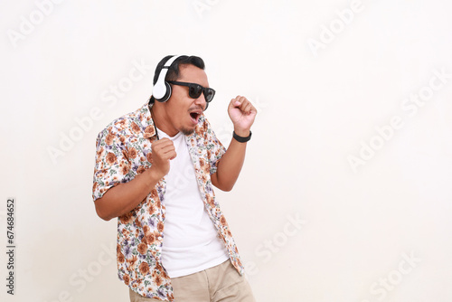 Happy Asian man traveler singing and listening music using headphone while dancing. Isolated on white background with copyspace photo