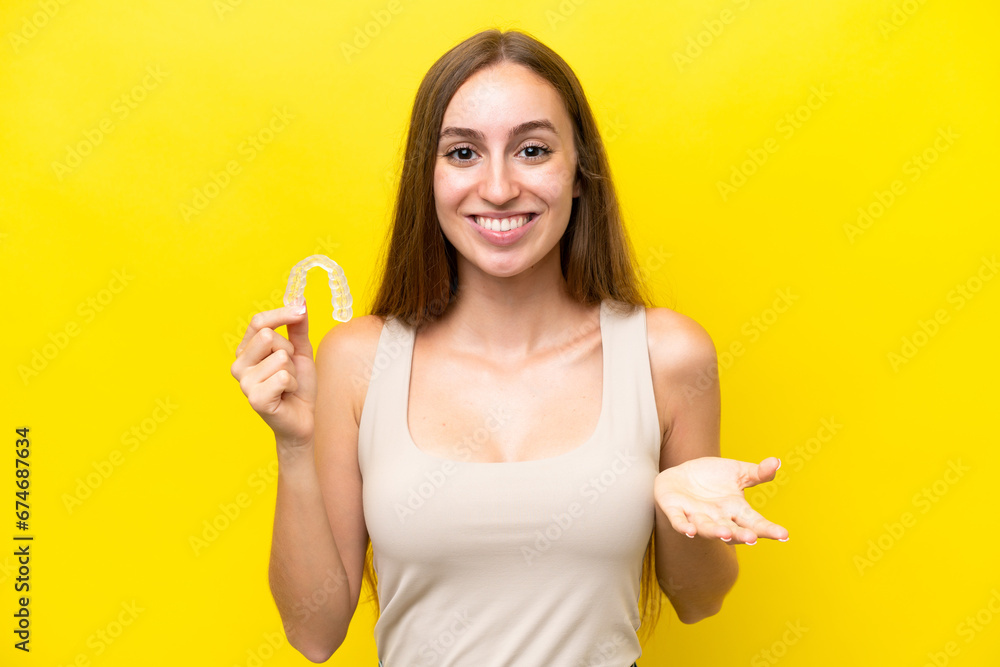 Young caucasian woman holding invisible braces isolated on yellow background with shocked facial expression Young caucasian woman holding invisible braces isolated on yellow background with shocked facial expression