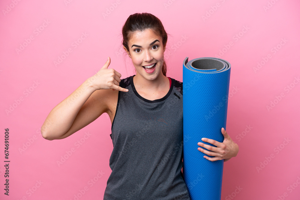 Young Brazilian sport woman going to yoga classes while holding a mat isolated on pink background making phone gesture. Call me back sign Young Brazilian sport woman going to yoga classes while holding a mat isolated on pink background making phone gesture. Call me back sign