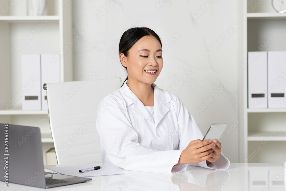 Smiling asian female doctor using smartphone while sitting at desk in clinic, Smiling asian female doctor using smartphone while sitting at desk in clinic,