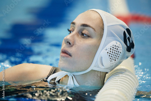 Young restful female water polo player in swimming cap standing in deep water of pool by finish line and having break between trainings photo