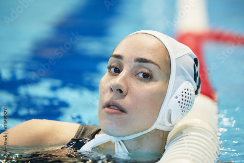 Face of young serene female swimmer in cap standing in deep water of swimming pool and looking at camera while having break photo