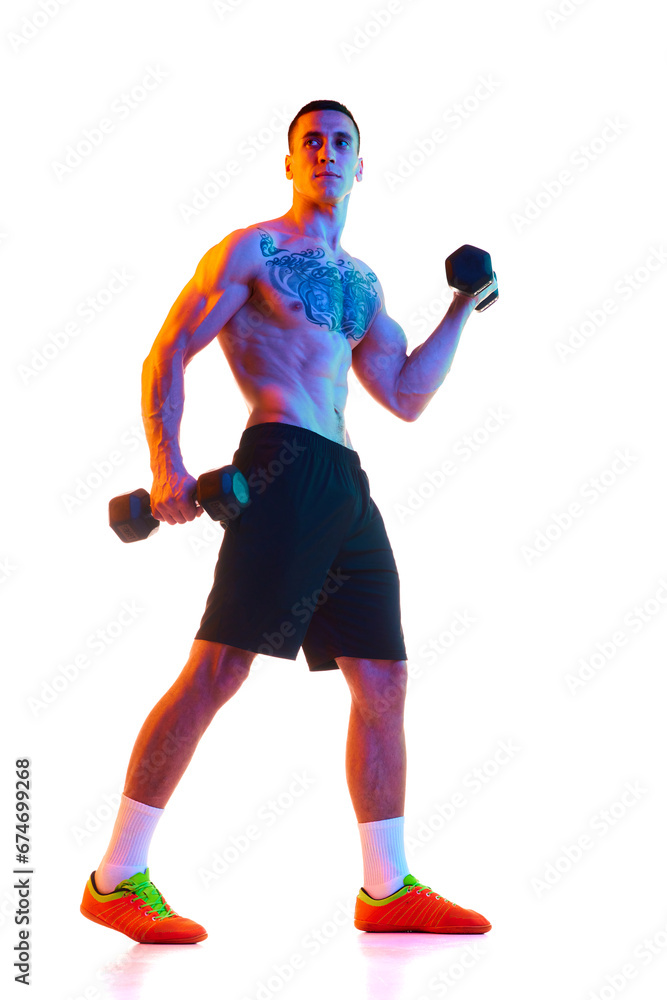 Full-length of shirtless young man with strong, muscular fit body training with dumbbells against white studio background in neon light. Concept of sport, active and healthy lifestyle, body, fitness