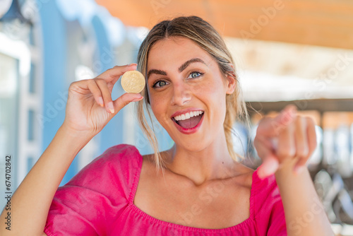 Young blonde woman holding a Bitcoin at outdoors points finger at you with a confident expression