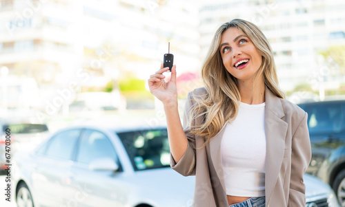 Pretty blonde Uruguayan woman holding car keys at outdoors looking up while smiling