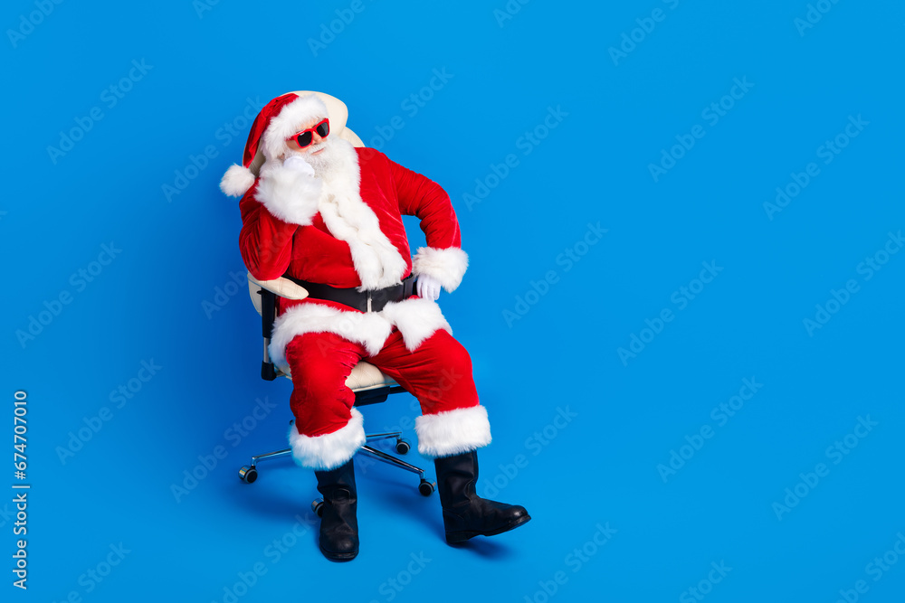 Full size photo of pensioner man armchair look empty space dressed stylish santa claus costume coat isolated on blue color background Full size photo of pensioner man armchair look empty space dressed stylish santa claus costume coat isolated on blue color background