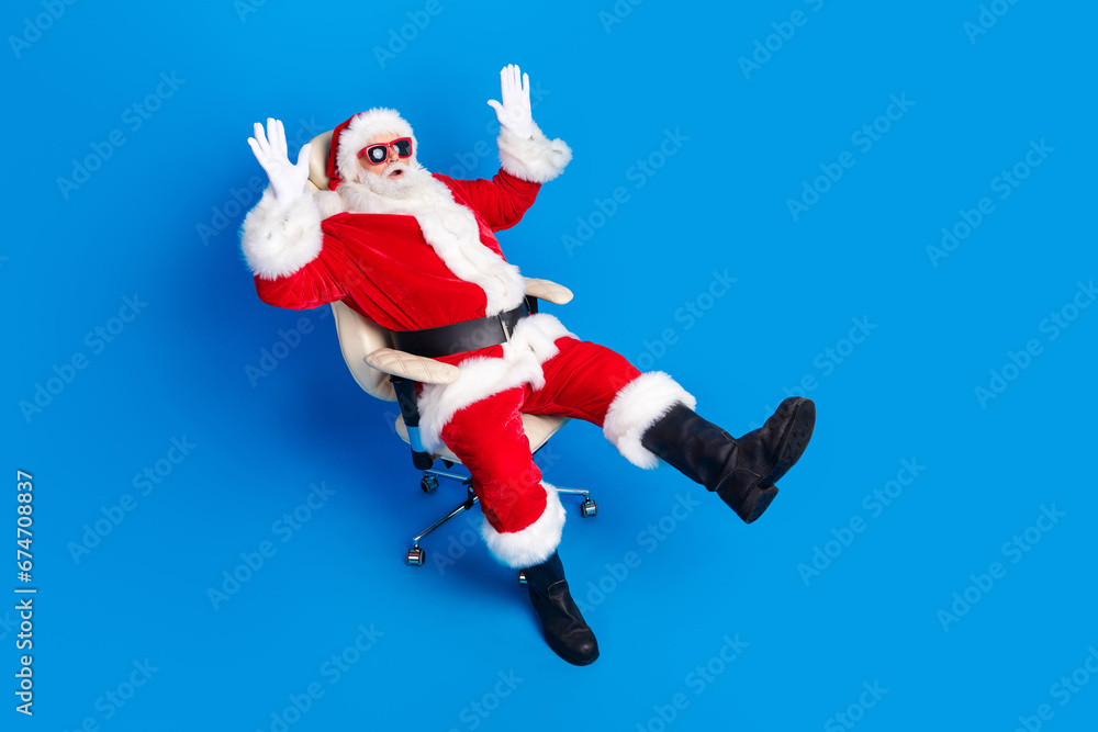 Full size photo of pensioner man astonished raise hands armchair dressed stylish santa claus costume coat isolated on blue color background Full size photo of pensioner man astonished raise hands armchair dressed stylish santa claus costume coat isolated on blue color background