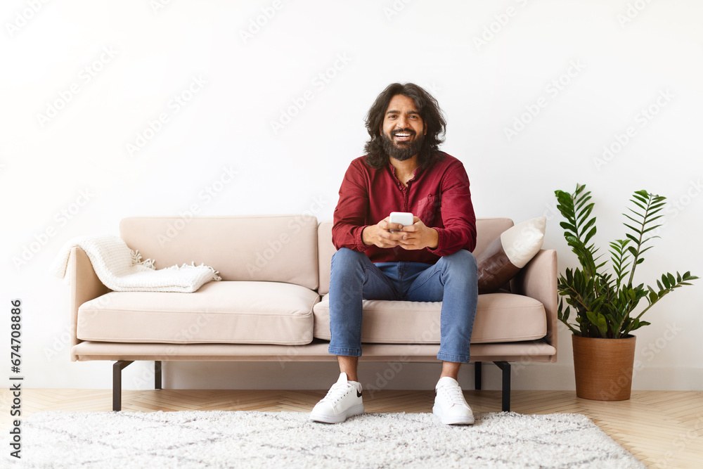 Positive indian man sitting on couch and using cell phone Positive indian man sitting on couch and using cell phone