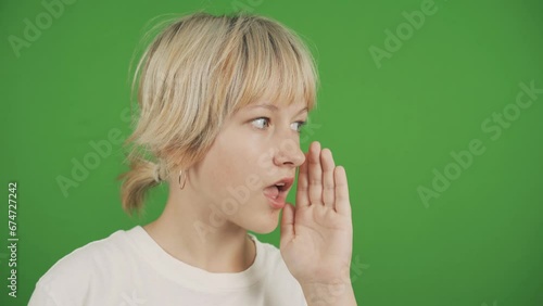 A teenage girl puts her hand to her mouth and quietly shares secret information. Close-up. Isolated on a green background, chromakey. photo