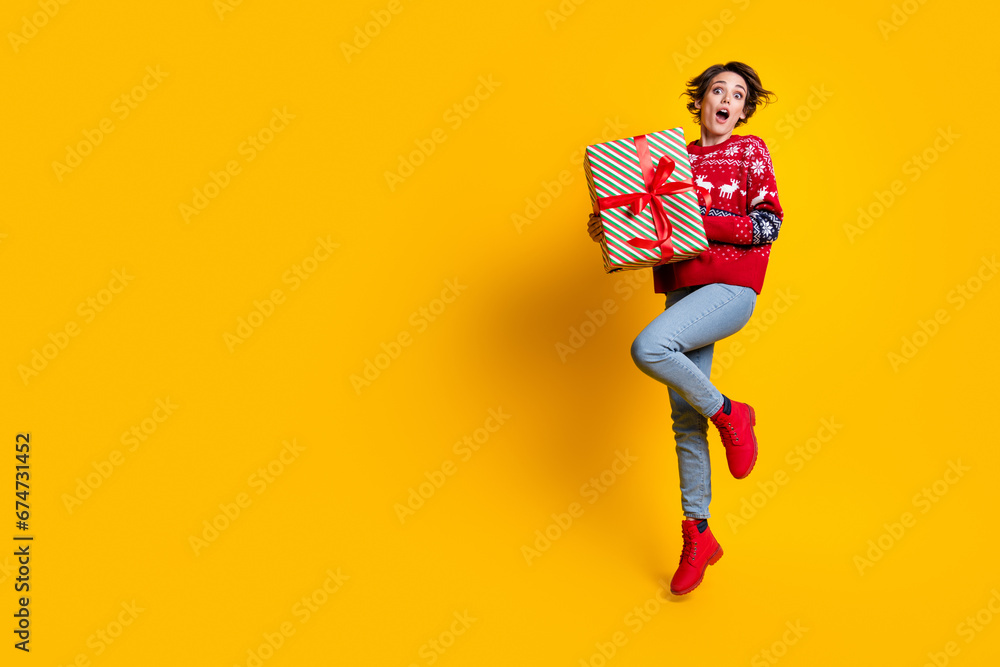 Fototapeta premium Full body photo of impressed astonished woman wear sweater hold present box jumping near empty space isolated on yellow color background