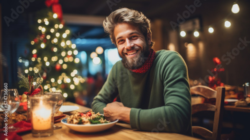Cheerful man with gray hair in a brown sweater, sitting at a festive Christmas dinner table with a glass of wine, enjoying the cozy ambiance created by soft lighting and holiday decorations.