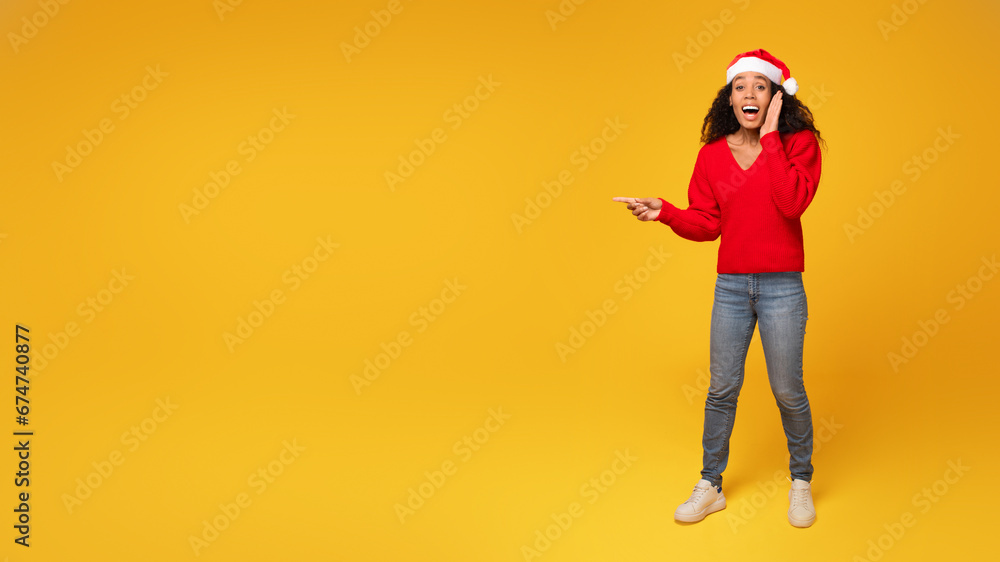 Surprised black lady in Santa hat points at space, yellow background Surprised black lady in Santa hat points at space, yellow background
