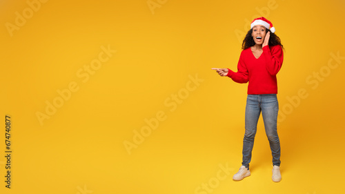 Surprised black lady in Santa hat points at space, yellow background