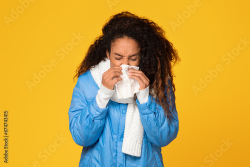 Ill black lady in jacket and scarf blows nose, yellow background