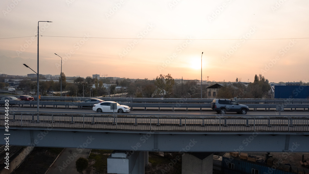 Drone view of the bridge passing through the city. Different cars pass by, from cars to trucks. Free lane. Against the background of a city at sunset in smog. The chimneys of the factory are smoking