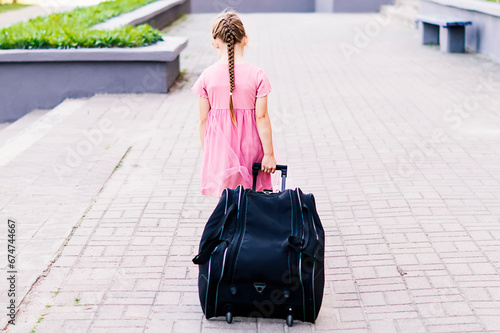 Rear view of a stylish little girl in a pink dress walking with luggage. Full length shot of a little girl walking and pulling a suitcase. A child rolls a suitcase on wheels along the sidewalk