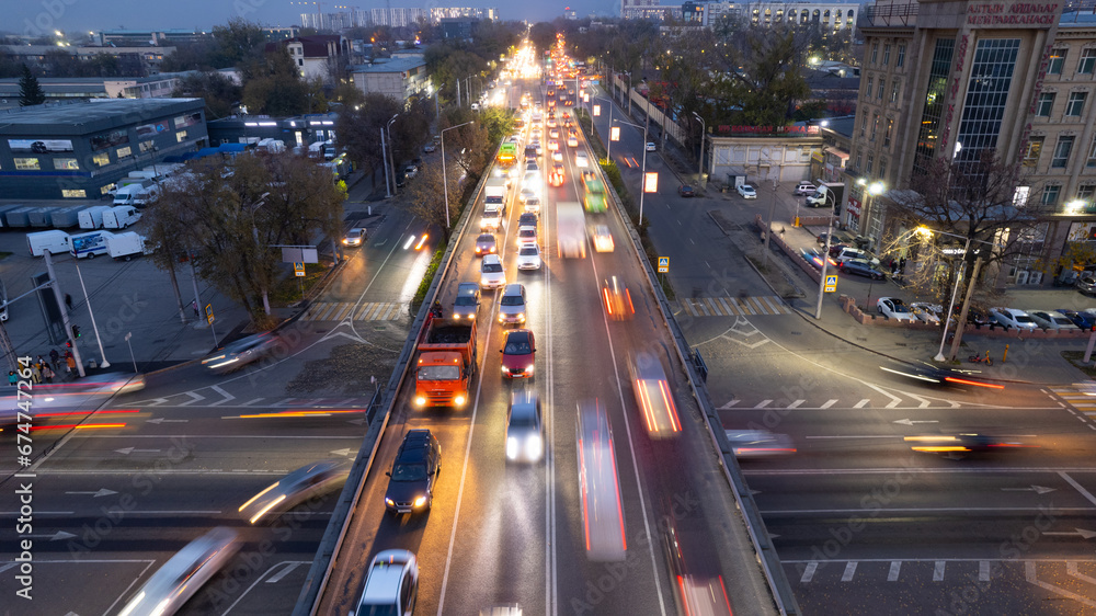 A busy intersection with a lot of cars, buses and trucks. Aerial view from the drone on the night road, the city and pedestrians. Lights and headlights are shining. A traffic jam has formed. Almaty