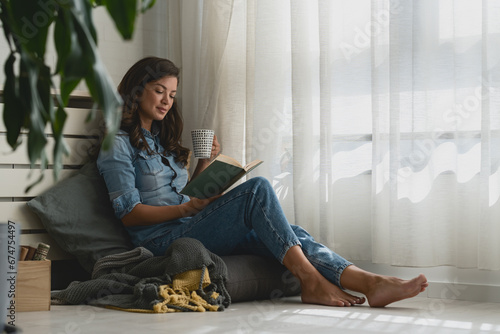 Handsome young woman reading a book, drinking coffee at home sitting cozy by the window