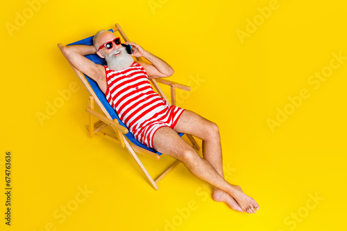 Full length portrait of carefree aged man sunbathing lounger speak telephone isolated on yellow color background