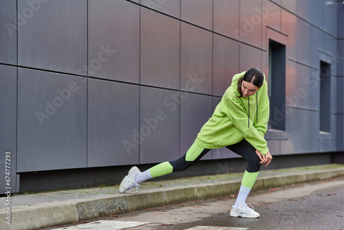 athletic woman in lime color hoodie and leggings doing lunges near grey wall outdoors, motivation