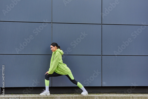 young sportswoman in hoodie and leggings stretching legs near grey wall outdoors, energy and sport