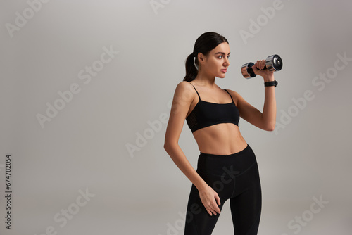 slim and strong young woman in active wear lifting dumbbell while working out on grey backdrop © LIGHTFIELD STUDIOS