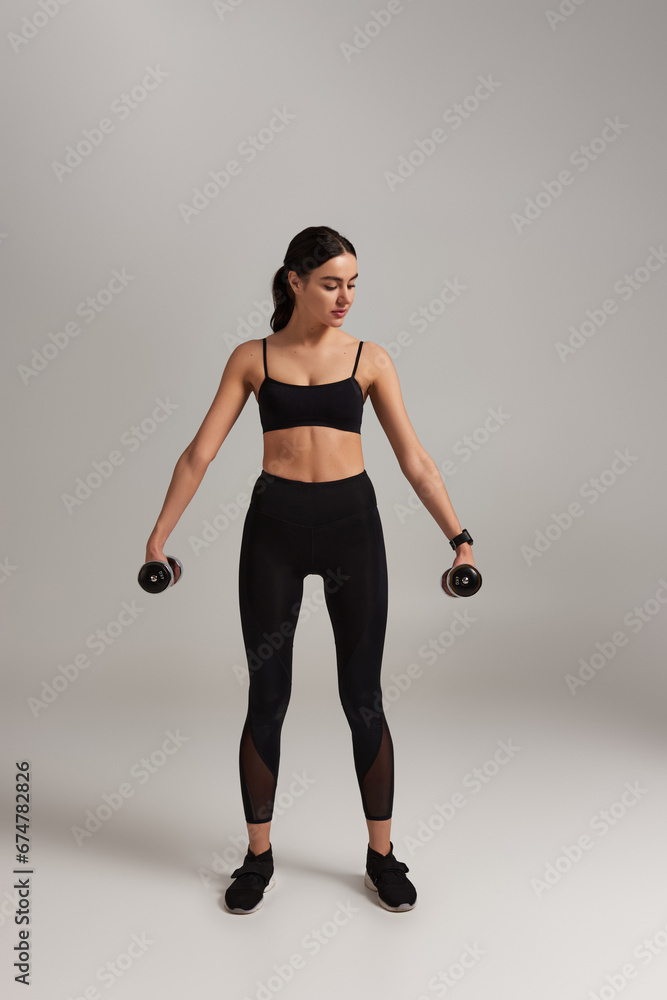 full length of pretty sportswoman in black active wear exercising with dumbbells on grey backdrop
