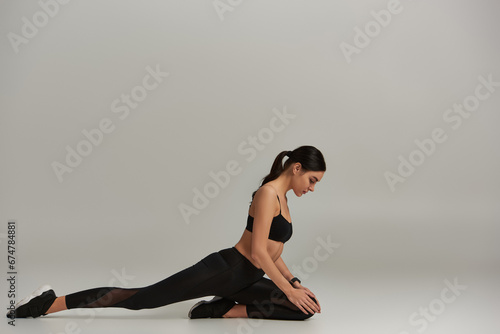 side view of flexible woman in active wear and sneakers stretching on grey background, motivation
