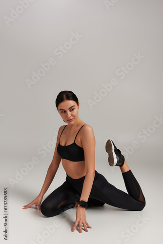flexible young sportswoman in black leggings and crop top stretching legs on grey backdrop