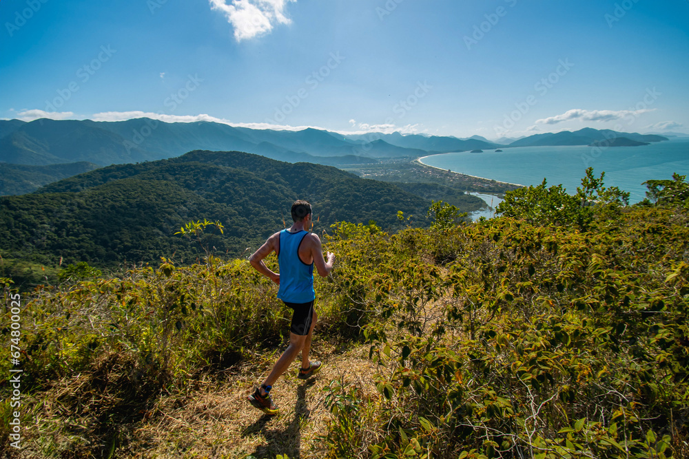 Corredor atleta de montanha correndo no topo do morro no meio da mata com vista para o oceano em dia de sol no verão