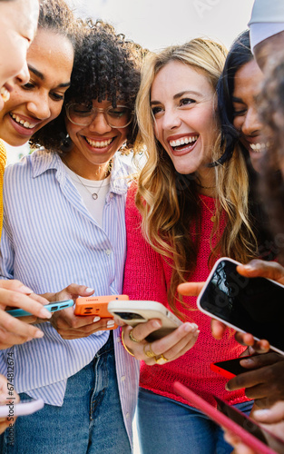 Happy female group using mobile phone together outdoors. Diverse women friends laughing while watching funny social media content on cellphone network app. Friendship and technology lifestyle concept