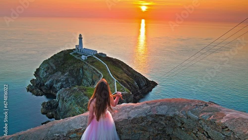 Beautiful woman in a dress visits South Stack Lighthouse at golden sunset in Holy Island, Wales. A tourist in a prom wedding dress walks at the Lighthouse with a footbridge over wild Atlantic ocean. photo