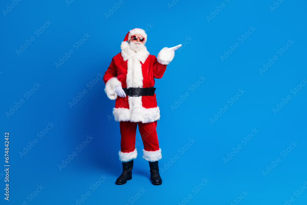 Full size photo of pensioner senior man palm hold empty space dressed stylish santa claus costume coat isolated on blue color background Full size photo of pensioner senior man palm hold empty space dressed stylish santa claus costume coat isolated on blue color background
