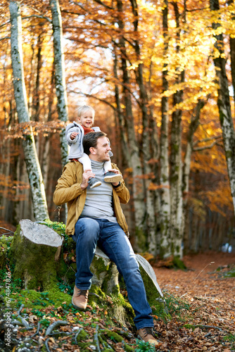 Little smiling girl sits on the shoulders of her dad sitting on a stump in an autumn park