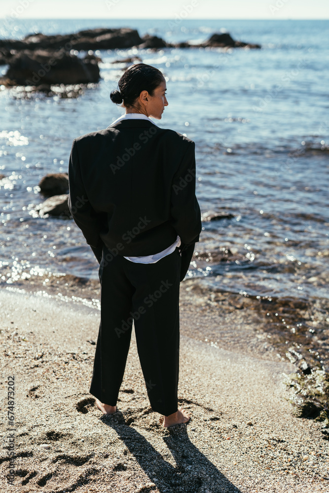 Brunette girl in a black men's suit barefoot on the beach by the sea. Fashion.