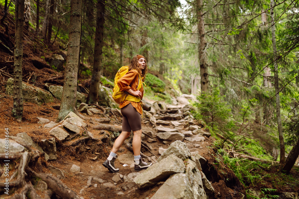 Obraz premium Smiling woman traveler along a forest hiking trail in the mountains against the backdrop of nature. Young woman with backpack traveling outdoors. Hiking, active lifestyle.