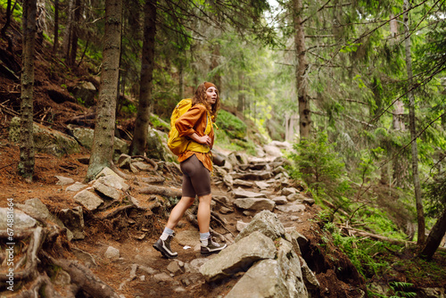 Smiling woman traveler along a forest hiking trail in the mountains against the backdrop of nature. Young woman with backpack traveling outdoors. Hiking, active lifestyle.