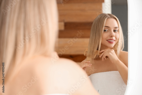 Young blonde woman combing hair near mirror in bathroom