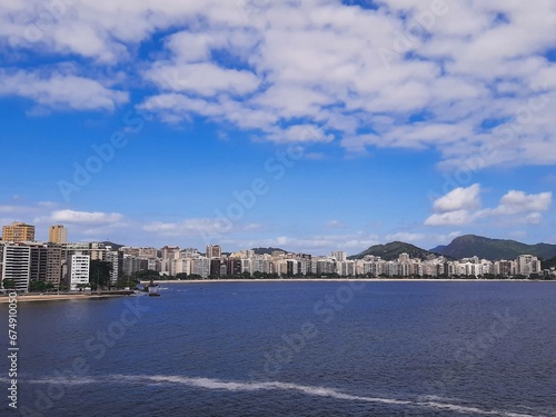 View of Icaraí Beach in Niterói, Rio de Janeiro, Brazil, tall buildings, calm waters of Guanabara Bay and blue sky with clouds on a spring afternoon © Hello_Brazil