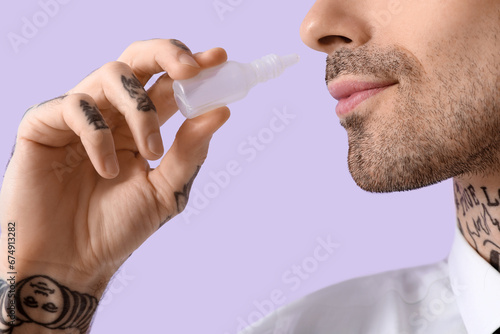 Fototapeta Naklejka Na Ścianę i Meble -  Allergic young man using nasal drops on lilac background, closeup
