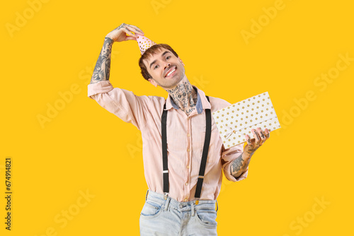 Happy young man with gift box and party hat on yellow background